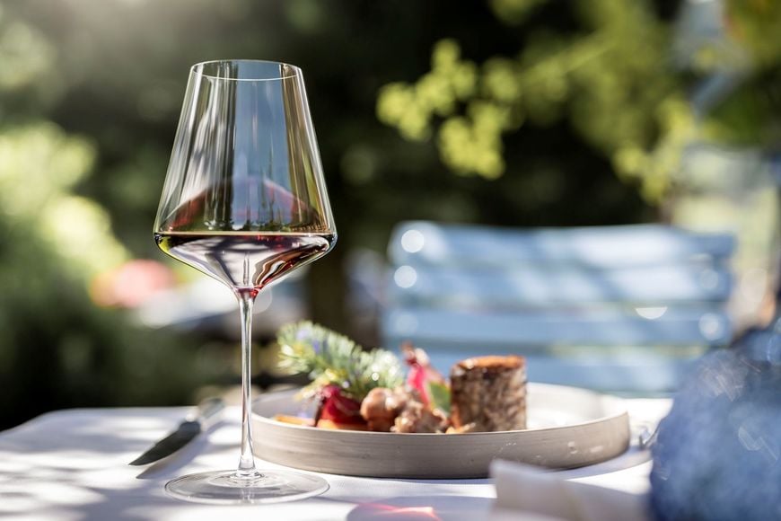 A glass of red wine on a set table outdoors, with a plated dish in the background.