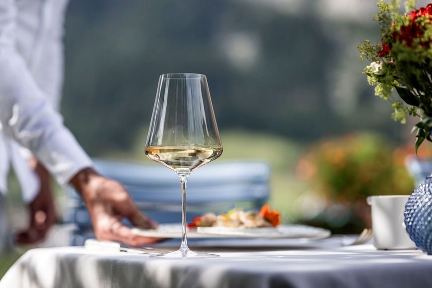 A glass of white wine on a set table, with a served dish on a plate in the background.