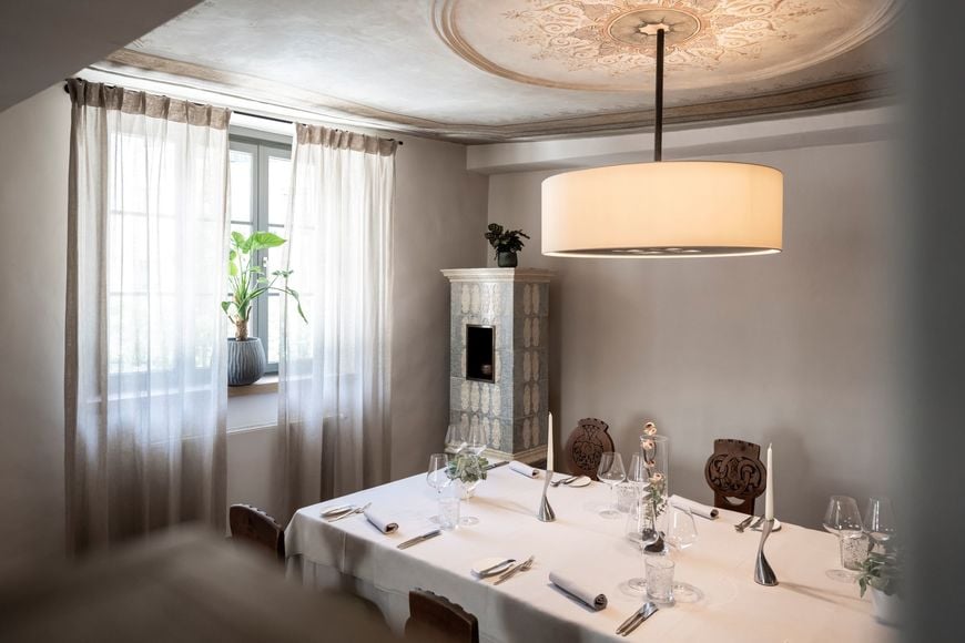 Set table at Restaurant Löwengrube with antique wooden chairs, plant by the window, designer lamp, and a tiled stove in the corner. ©Löwengrube/Alex Filz