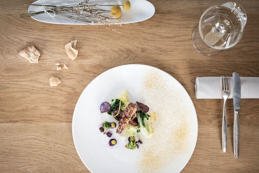 Fish dish on wooden table with white wine glass, Schüttelbrot, cutlery, and floral decoration. ©Löwengrube/Alex Filz