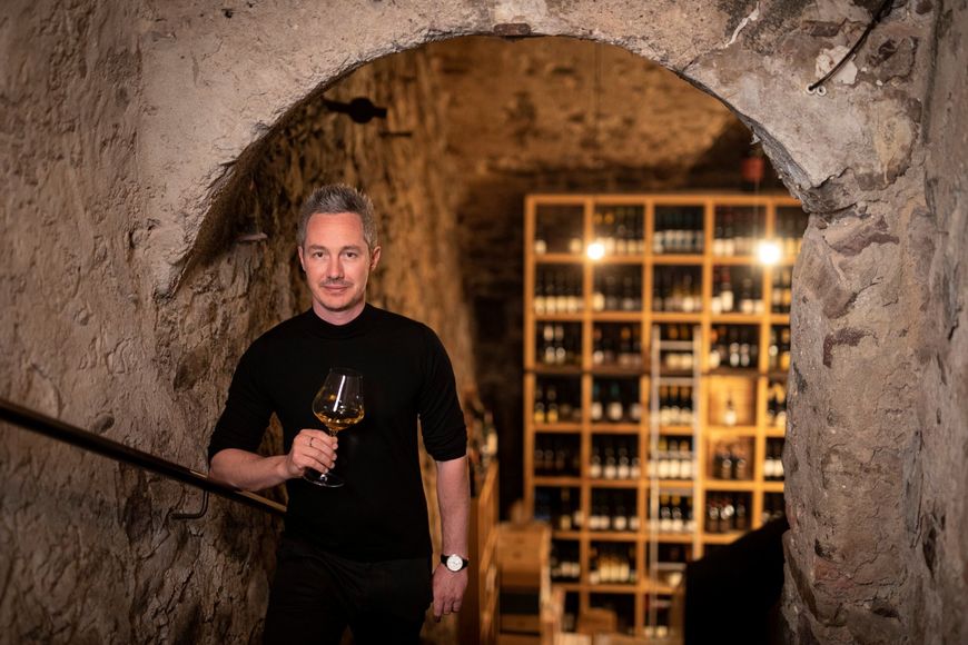 Michael Meister from Restaurant Löwengrube holding a white wine glass on the cellar stairs in front of the wine cellar. ©IDM/Consorzio Vini Alto Adige//Marion Lafolger