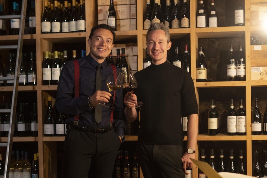 Two people clinking glasses of red wine in front of a wine rack, smiling at the camera. ©IDM/Consorzio Vini Alto Adige//Marion Lafolger