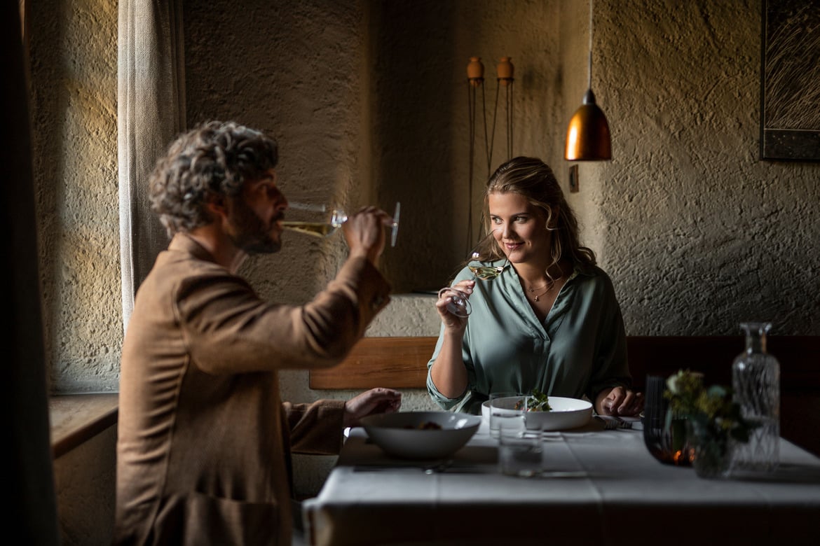 A couple sitting at a set table; the man drinks white wine, the woman smells a glass of white wine.